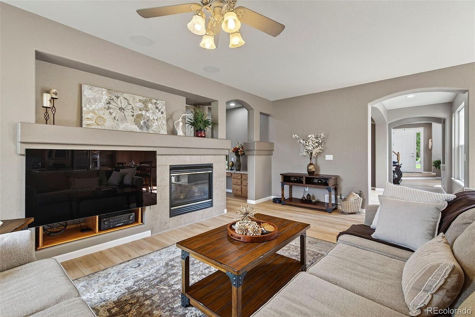 This is a warm and inviting living room featuring a fireplace with a mounted television, a comfortable sectional sofa, and a stylish coffee table. The room is decorated in neutral tones with hardwood floors and an area rug, creating a cozy and elegant atmosphere. An arched doorway leads to another room, enhancing the open feel of the home.