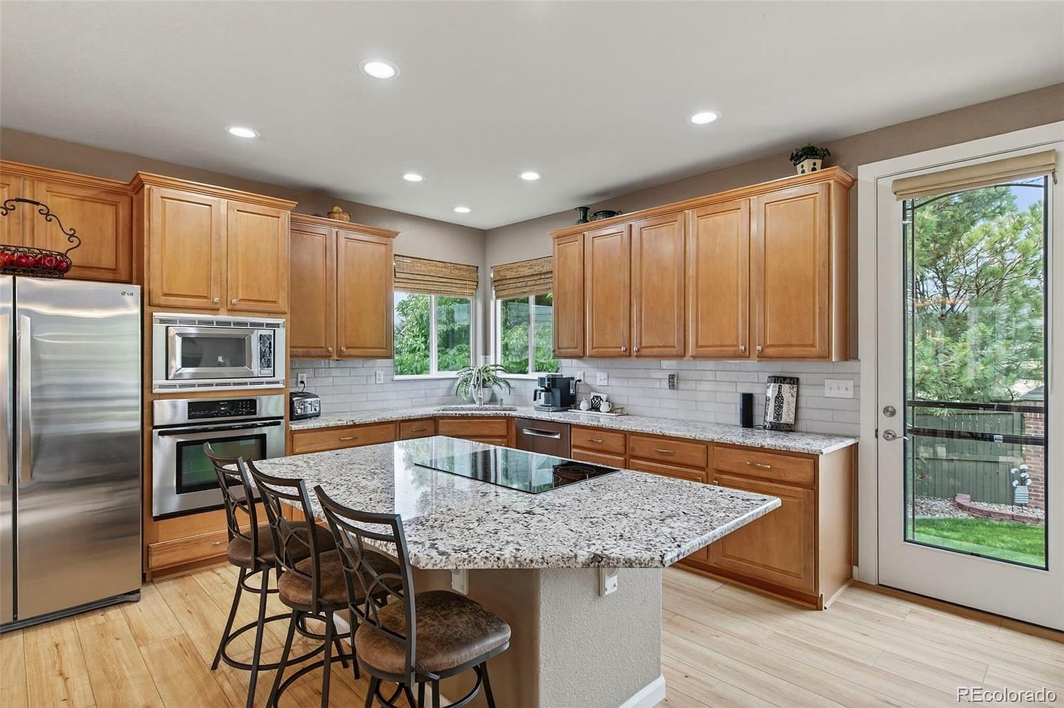 This is a well-lit kitchen featuring wooden cabinetry, stainless steel appliances, and a granite-topped island with bar seating. The kitchen has a neutral color palette with light wood flooring and a window providing natural light. The layout is spacious and functional, ideal for cooking and entertaining.