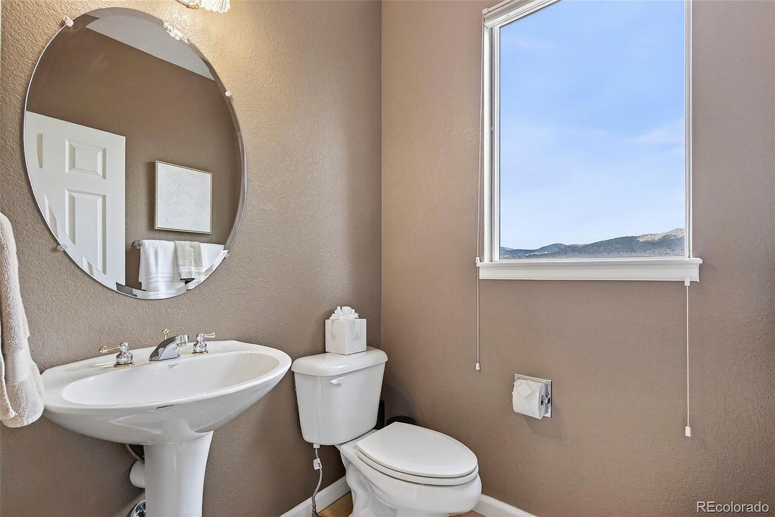 This is a well-lit guest bathroom featuring a pedestal sink with chrome fixtures, an oval mirror, and a standard toilet with a tissue box on top. A window provides natural light and a view of the outside. The walls are painted in a neutral tone, creating a clean and inviting space.
