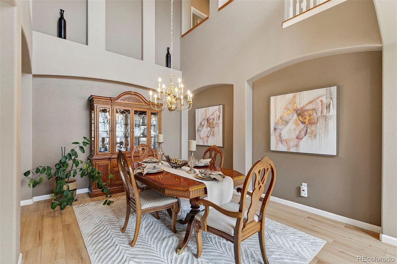 This is an interior shot of a dining room featuring a wooden dining table set with plates and silverware, surrounded by ornate wooden chairs. A large wooden china cabinet stands against the wall, and two framed abstract paintings hang on the adjacent wall. The room is illuminated by a chandelier, and a patterned rug covers the hardwood floor, creating a warm and inviting atmosphere.