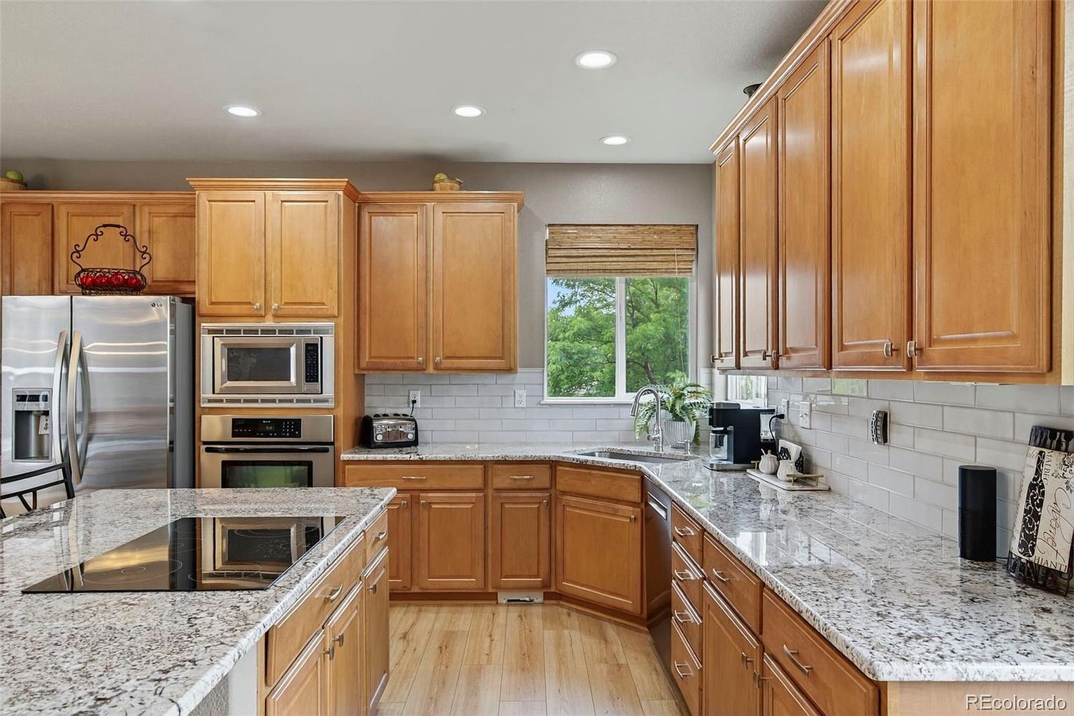 This is a well-lit kitchen featuring light wood cabinetry, granite countertops, and stainless steel appliances. The kitchen includes a central island with a cooktop, a built-in microwave and oven, and a window above the sink providing natural light. The overall impression is a clean and functional space.