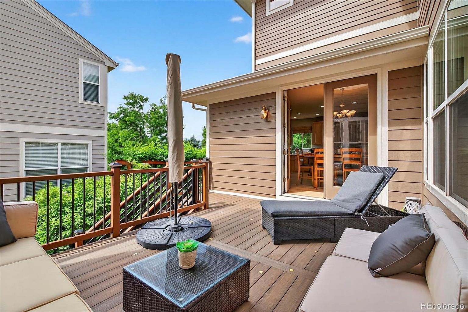 This image showcases a well-appointed outdoor deck area, perfect for relaxation and entertaining. The deck features comfortable lounge furniture, including a chaise lounge and a sofa with pillows. A glass-topped coffee table and a closed umbrella add to the ambiance, while sliding glass doors provide easy access to the interior of the home. The deck is surrounded by a railing, offering a sense of privacy and safety.