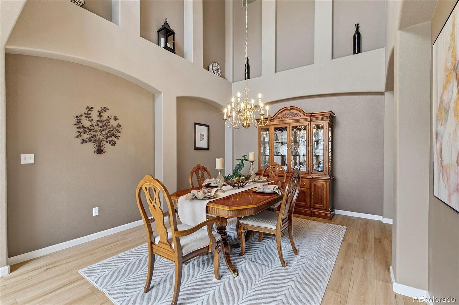 This is an interior shot of a dining room featuring a wooden dining table set with plates and a runner, surrounded by four chairs. A wooden china cabinet stands against the wall, and a chandelier hangs above the table. The room is decorated with neutral tones and features architectural niches with decorative items, creating a warm and inviting atmosphere.