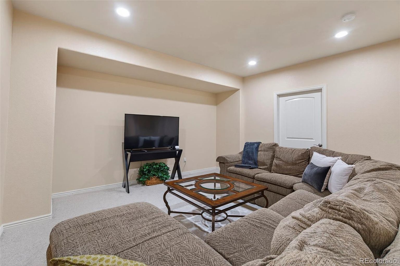This is an interior shot of a living room featuring a large, comfortable-looking sectional sofa in a neutral brown tone. A glass-topped coffee table sits in the center of the room, and a television is positioned against the wall on a modern stand, with a decorative plant underneath. The room is well-lit with recessed lighting, creating a cozy and inviting atmosphere.