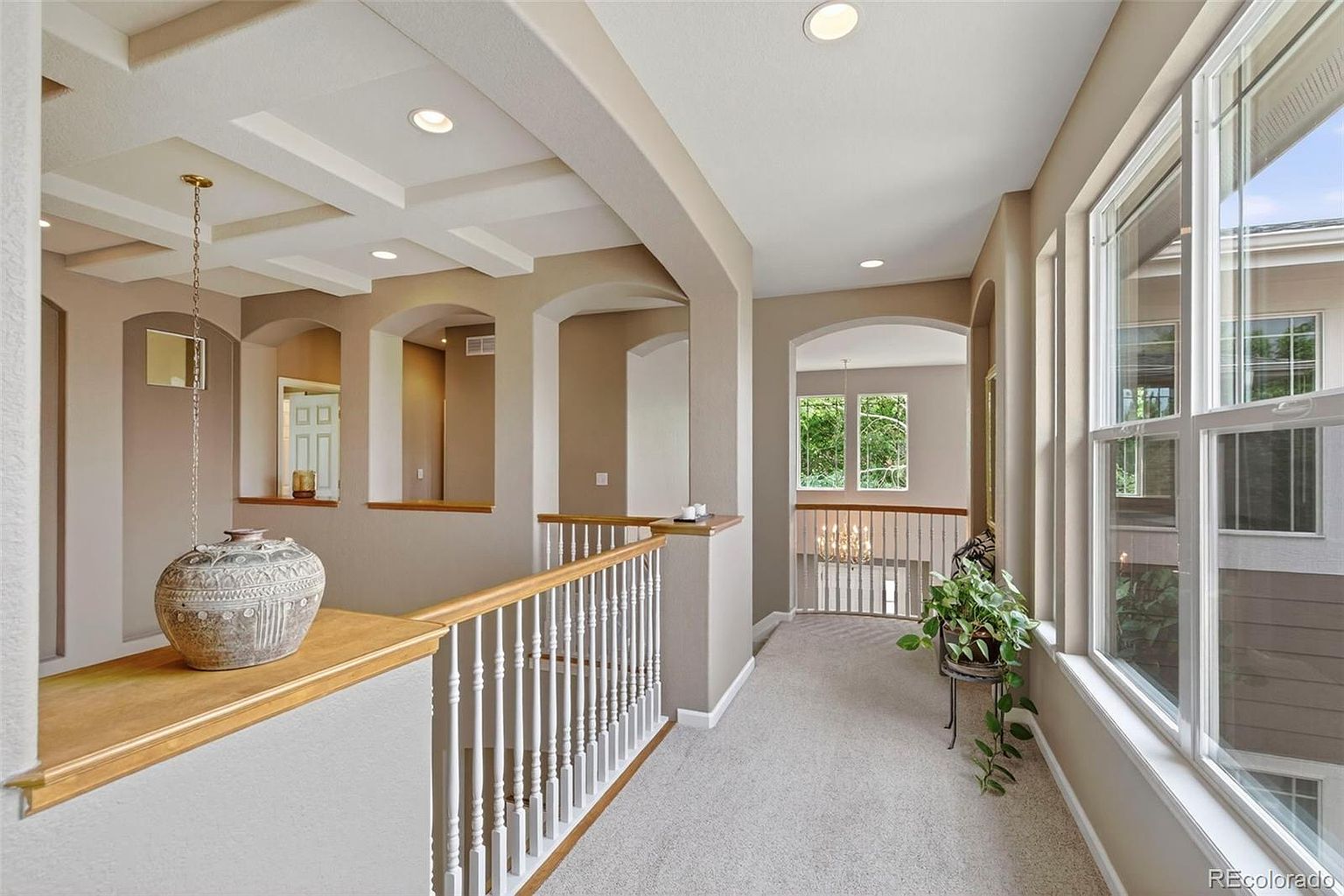 This interior shot showcases a well-lit hallway with architectural details such as arched openings and a coffered ceiling. The hallway features neutral-toned walls, carpeted flooring, and a wooden railing with white spindles. Natural light streams in through large windows, enhancing the bright and airy feel of the space.