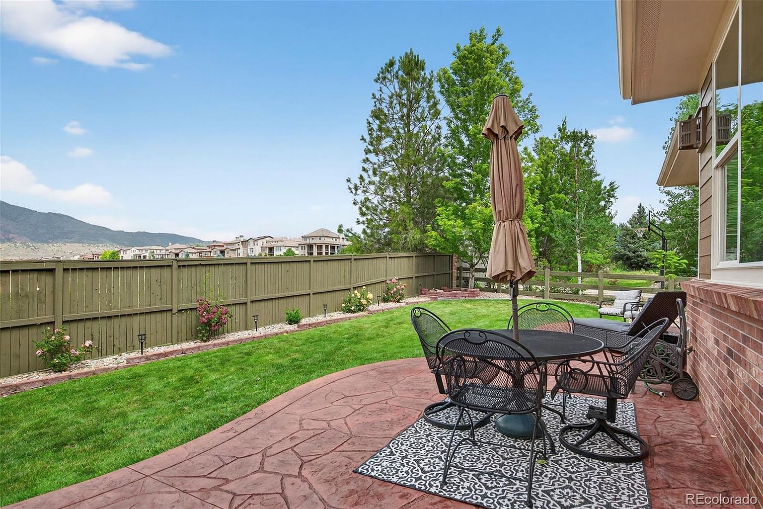This is a rear exterior view showcasing a well-maintained patio and backyard. The patio features a stone surface with an outdoor dining set, umbrella, and a decorative rug. The backyard includes a green lawn, a wooden fence with flower beds, and mature trees in the background, creating a private and inviting outdoor space.