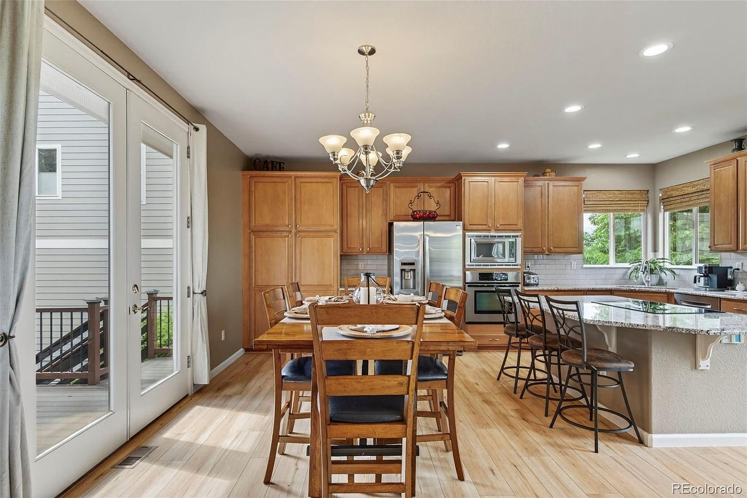 This interior shot showcases a well-lit kitchen and dining area. The kitchen features wooden cabinetry, stainless steel appliances, and a granite countertop island with bar seating. The dining area includes a wooden table set for a meal, complemented by a chandelier and a view to the outside through glass doors, creating a warm and inviting atmosphere.