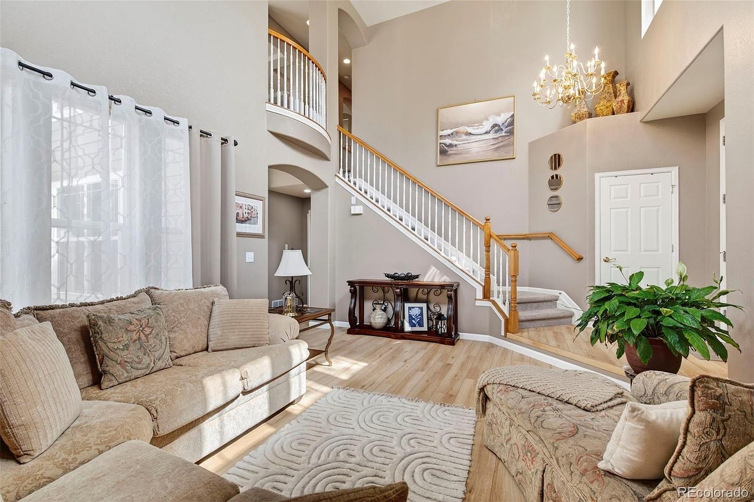 This is an inviting living room featuring a comfortable beige sofa with decorative pillows, a patterned armchair, and a light-colored area rug. A staircase with white railings and wooden banisters leads to the upper level, while a console table sits beneath the stairs. The room is well-lit with natural light and a chandelier, creating a warm and welcoming atmosphere.