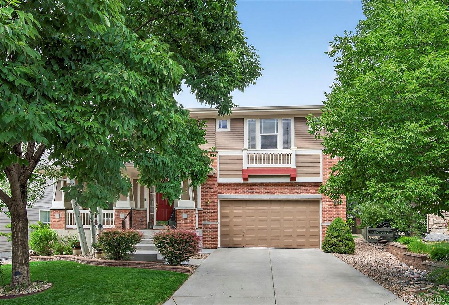 This is a front view of a two-story house with a brick and siding exterior. The house features a two-car garage, a small balcony above the garage, and a red front door. Landscaping includes a well-maintained lawn, bushes, and trees, creating a welcoming curb appeal.