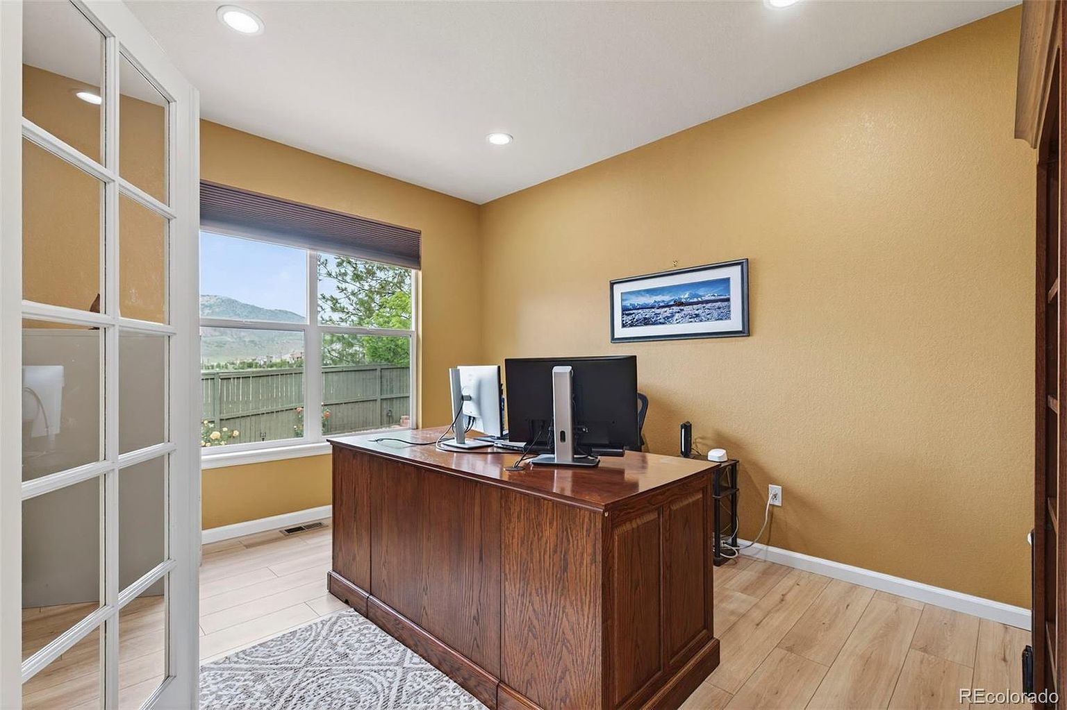 This is an interior shot of a home office featuring a large, wooden desk with computer monitors. The room has warm, yellow walls, light wood flooring, and a window offering a view of the outdoors. A framed picture hangs on the wall, and the overall impression is a professional and comfortable workspace.