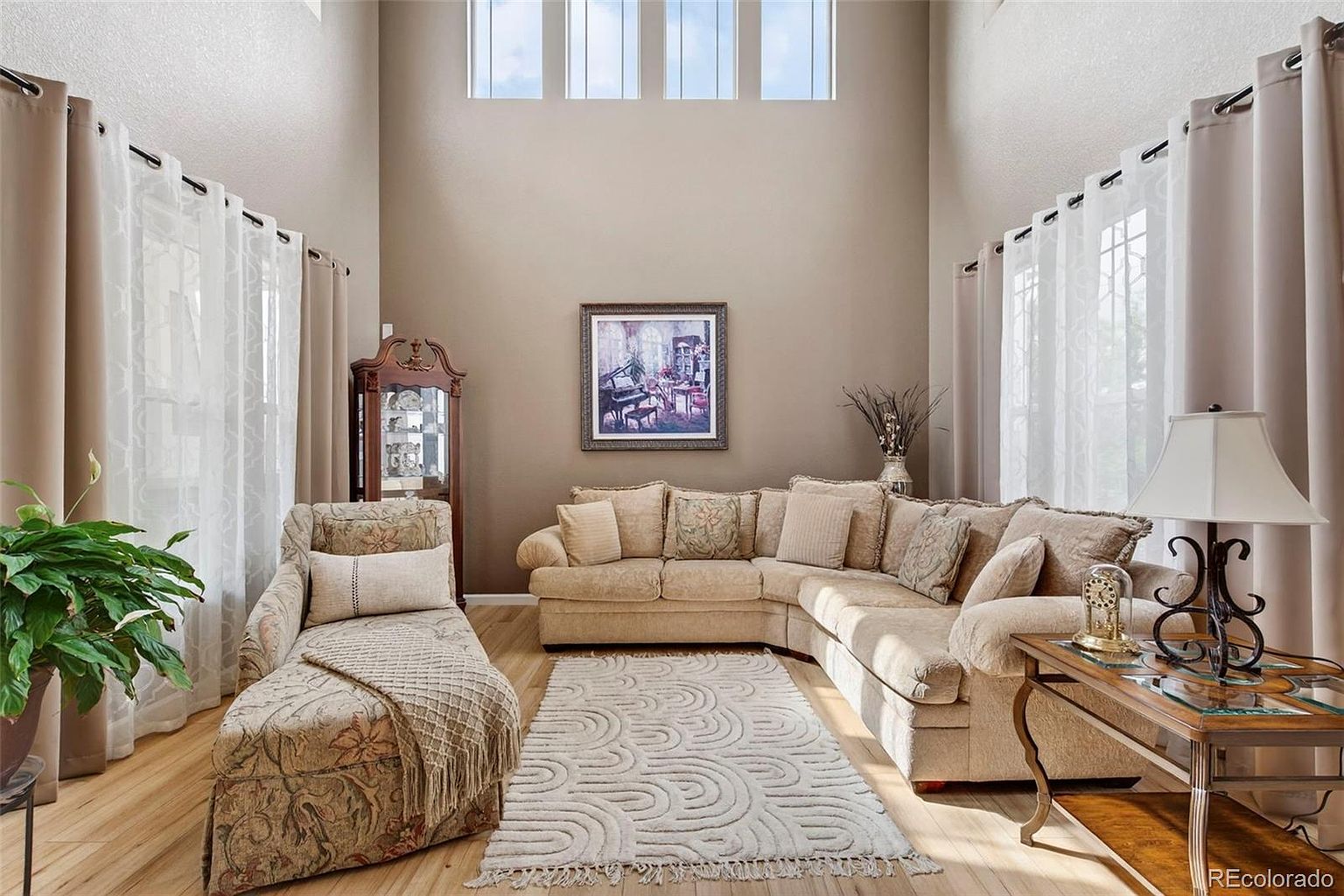 This is an interior shot of a living room featuring a large sectional sofa, a chaise lounge, and a decorative rug. The room has high ceilings with tall windows and neutral-toned walls, creating a bright and airy atmosphere. A curio cabinet and a framed painting add to the room's traditional style.