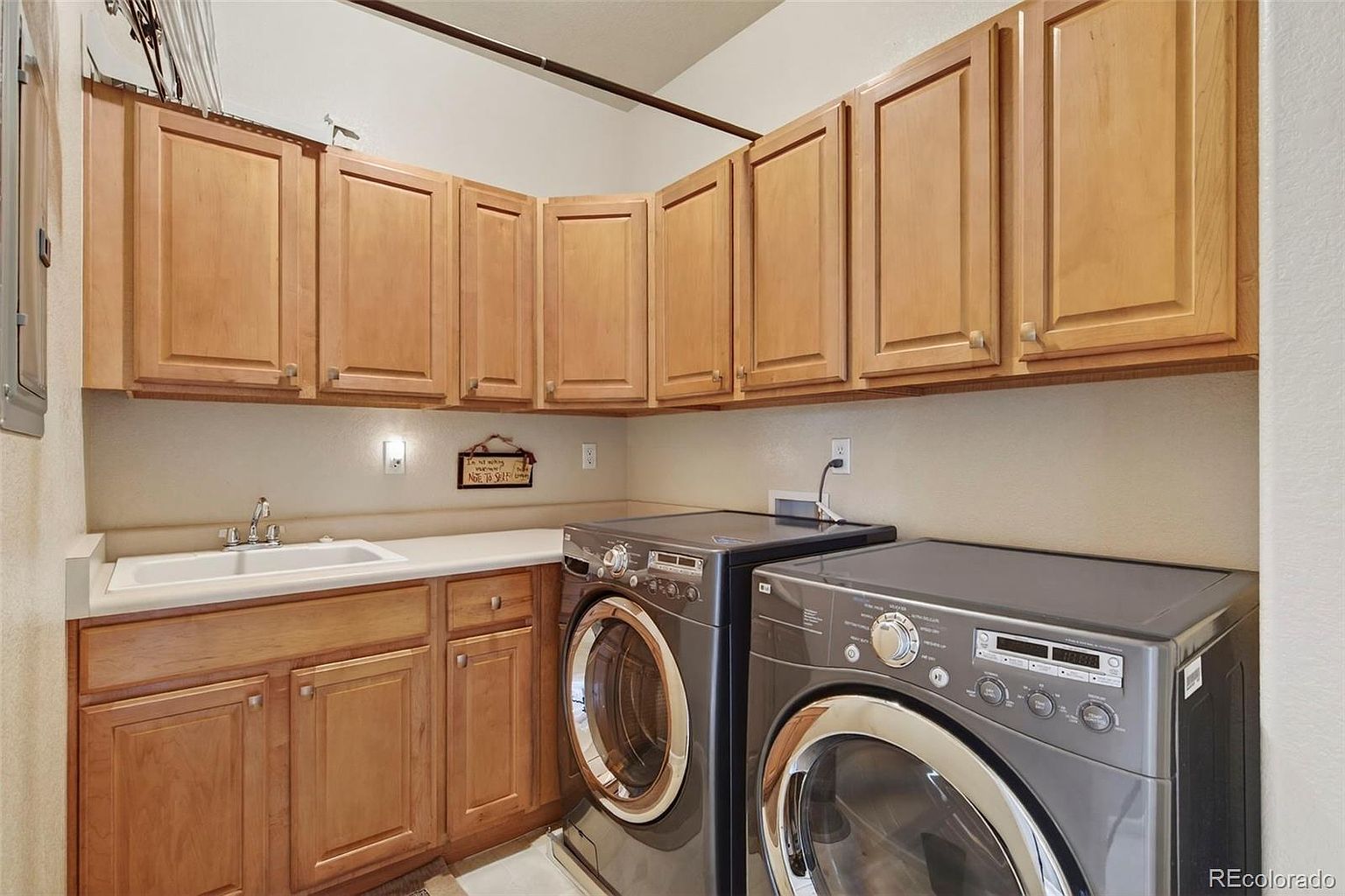 This is an interior shot of a laundry room featuring light wood cabinets above and below a white countertop with a sink. A gray front-loading washer and dryer are positioned next to the cabinets. The room is well-lit and appears functional, offering a practical space for laundry tasks.