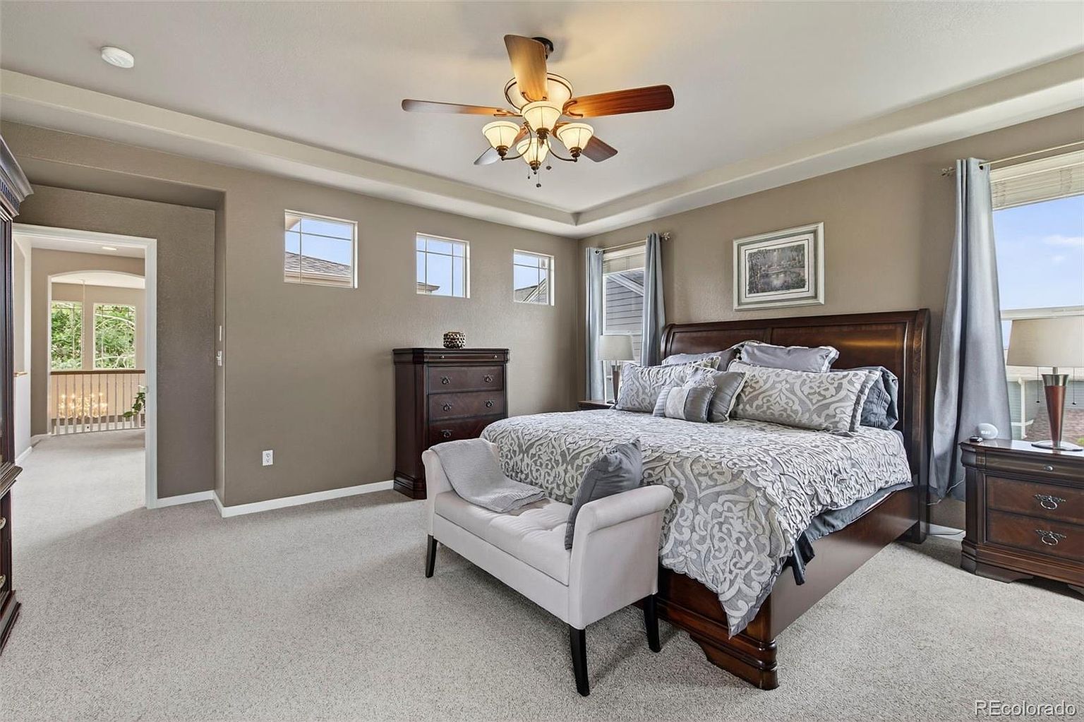 This is a primary bedroom featuring a large bed with a patterned comforter and several pillows. The room has a neutral color palette with taupe walls and light gray carpet. There is a ceiling fan with lights, a dresser, and a bench at the foot of the bed. The room has a calm and inviting atmosphere.