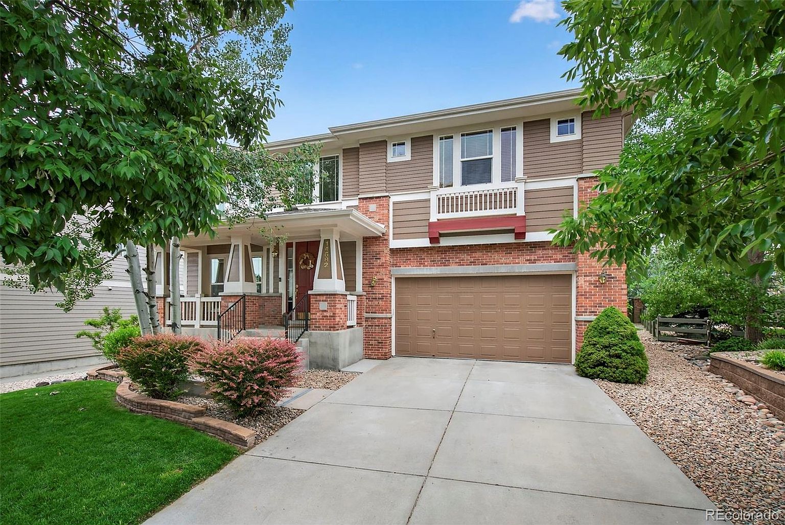 This is a front view of a two-story house with a brick and siding exterior. The house features a covered front porch with decorative columns, a two-car garage, and well-maintained landscaping. The overall impression is a welcoming and well-cared-for family home.
