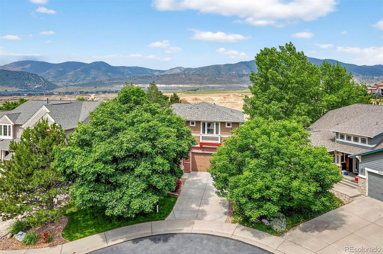 This aerial view showcases a well-maintained suburban home nestled among mature trees. The property features a neatly manicured lawn, a paved driveway leading to an attached garage, and a balcony. In the background, rolling hills and a partly cloudy sky add to the scenic appeal, suggesting a peaceful and desirable location.