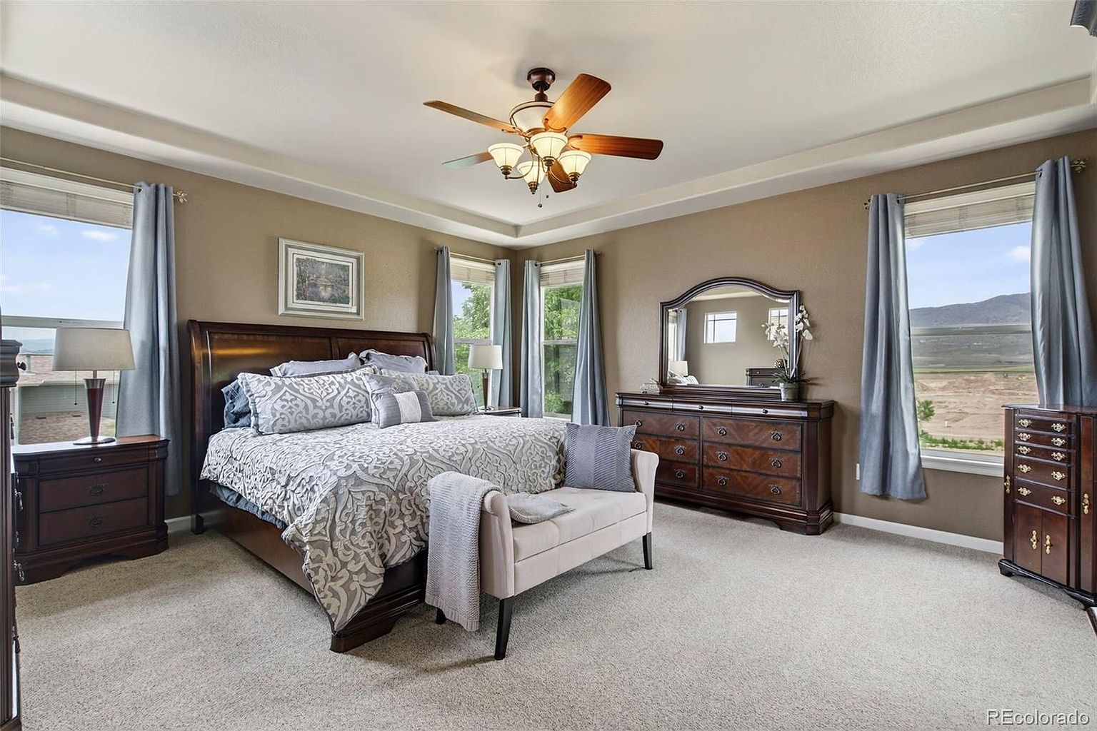 This is a well-appointed primary bedroom featuring a large bed with decorative pillows and a patterned comforter. The room has a neutral color palette with brown walls and light carpeting. Natural light streams in through the windows, which are framed by blue curtains. A ceiling fan with lights is centered in the room, and there is a dresser with a mirror and a bench at the foot of the bed.