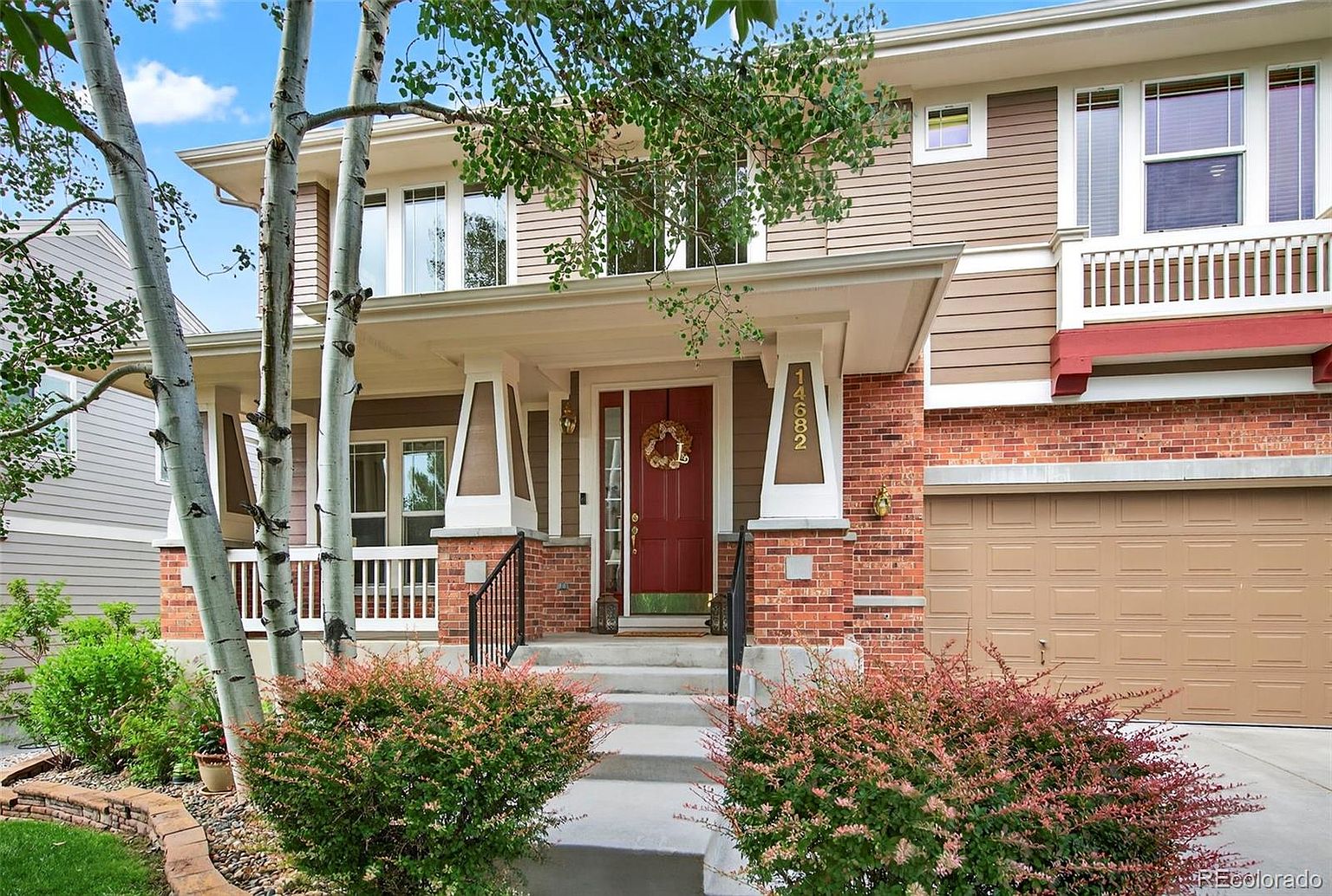 This is a front view of a two-story house with a brick and siding exterior. The house features a covered front porch with white columns and a red front door adorned with a wreath. Landscaping includes bushes and trees, adding to the curb appeal. The house number '14682' is prominently displayed on one of the porch columns.