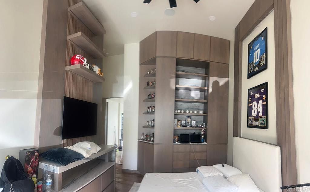 This bedroom features custom wood-toned cabinetry and shelving units that provide ample storage and display space for sports memorabilia. A wall-mounted television is positioned above a low-profile console, while two framed sports jerseys are displayed on the wall above a white bed. The room has a modern, organized aesthetic with a neutral color palette and integrated lighting.