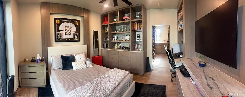 This contemporary bedroom features a central bed framed by a custom light wood built-in unit that includes open shelving and a display area for a framed sports jersey. To the right, a long, wall-mounted desk provides a dedicated workspace beneath a large, wall-mounted television. The room is finished with light wood-toned flooring and leads into a walk-in closet, creating a cohesive and functional living space.