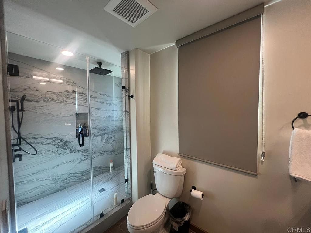 This modern bathroom features a walk-in shower with large-format patterned tile walls and a rainfall showerhead. Adjacent to the shower is a standard white toilet and a neutral-toned wall with a roller shade covering the window. The space is clean and functional, with a towel ring mounted on the wall and a small wastebasket tucked beside the toilet.
