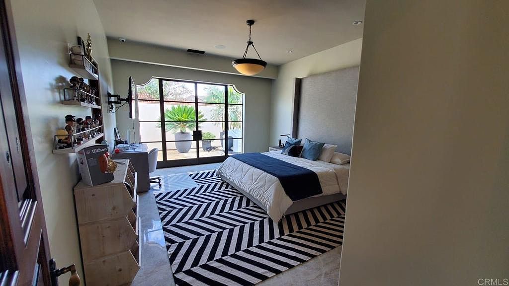 This spacious bedroom features a large bed with a tall, upholstered headboard and a bold, black-and-white chevron-patterned area rug. To the left, a light wood storage unit and a desk area are positioned near large glass doors that open to an outdoor patio. The room is illuminated by a central pendant light and natural light streaming through the glass doors, creating a bright and contemporary atmosphere.