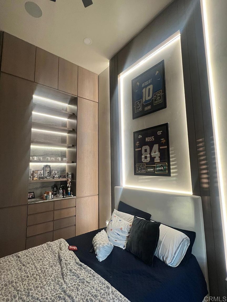 This modern bedroom features a custom light wood built-in storage unit with integrated shelving and recessed lighting. The focal point is a wall-mounted display of two framed sports jerseys, highlighted by vertical LED accent lighting. A bed with a white headboard and dark bedding sits in the foreground, creating a sleek and contemporary aesthetic.