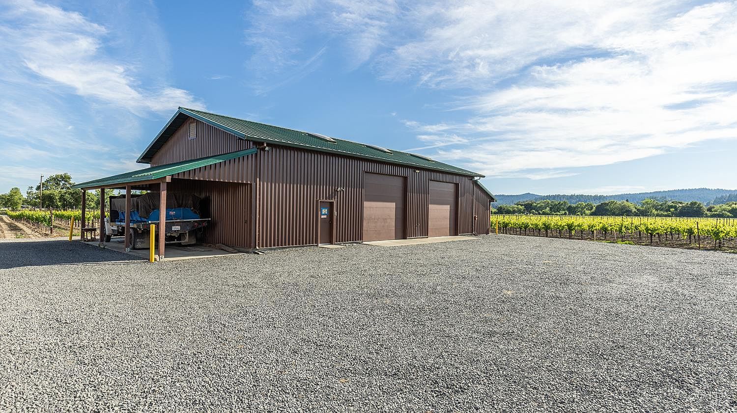 This image captures a large, brown metal-sided agricultural building set against a vast, gravel-covered lot. The structure features a covered side bay housing a truck, two large roll-up garage doors, and a green metal roof, all surrounded by expansive rows of grapevines under a bright, clear sky. The perspective is a wide-angle shot from the gravel lot, emphasizing the scale of the facility and its rural, vineyard-adjacent setting.