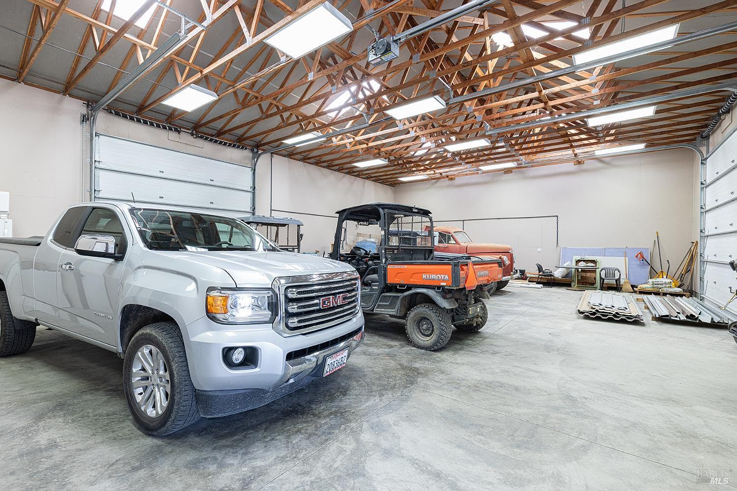 This spacious, high-ceilinged garage features exposed wooden trusses and multiple overhead light fixtures, providing ample room for vehicle storage. The concrete floor houses a silver pickup truck, an orange utility vehicle, and a vintage red truck parked in the background. The industrial-style space is equipped with large roll-up doors, making it a versatile area for automotive work or equipment storage.