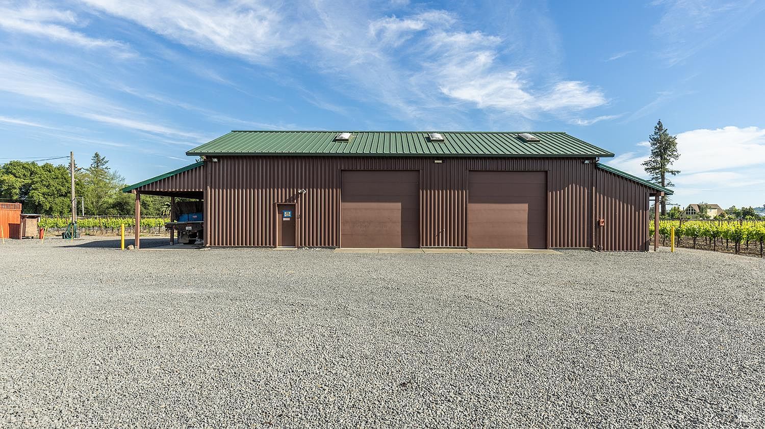 This image showcases a large, industrial-style metal barn or workshop set against a clear blue sky. The structure features a green corrugated metal roof, brown vertical siding, two large roll-up garage doors, and a side-access door, all situated on a wide gravel lot. The property is bordered by rows of grapevines, suggesting a rural or vineyard setting, and the perspective is a straight-on, eye-level shot that emphasizes the building's functional design.