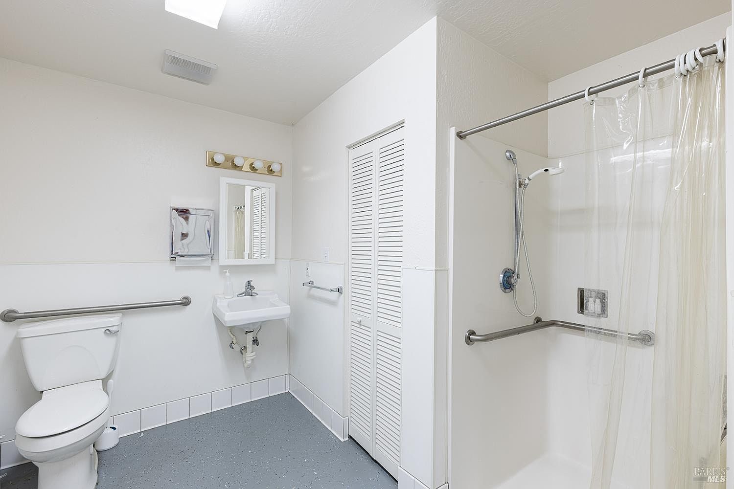 This accessible bathroom features a white toilet with a grab bar, a wall-mounted sink, and a walk-in shower equipped with a handheld showerhead and safety rails. The room has white walls, a grey floor, and a louvered closet door, creating a functional and utilitarian aesthetic. The perspective is a wide-angle shot capturing the entire layout of the accessible facilities.