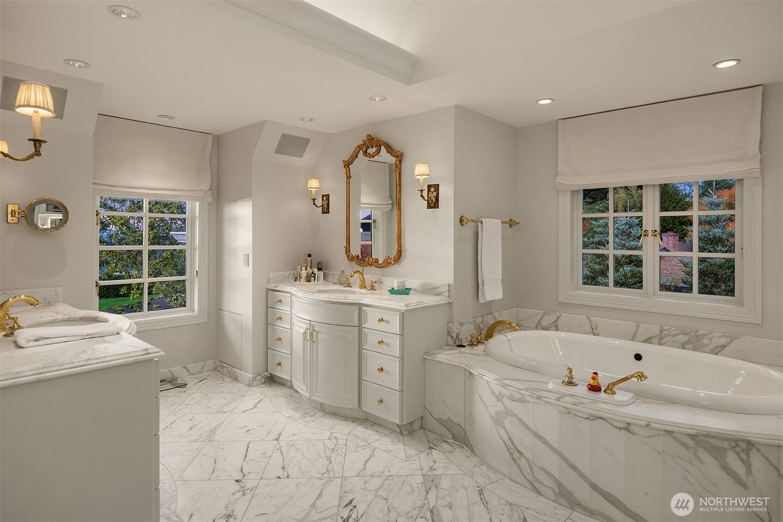 This luxurious primary bathroom features elegant marble flooring and countertops, a double vanity with gold-toned fixtures, and a deep soaking tub set against a marble-clad surround. The space is bright and airy, enhanced by large windows with soft Roman shades and a sophisticated gold-framed mirror. The overall aesthetic is classic, refined, and spa-like, offering a serene retreat within the home.