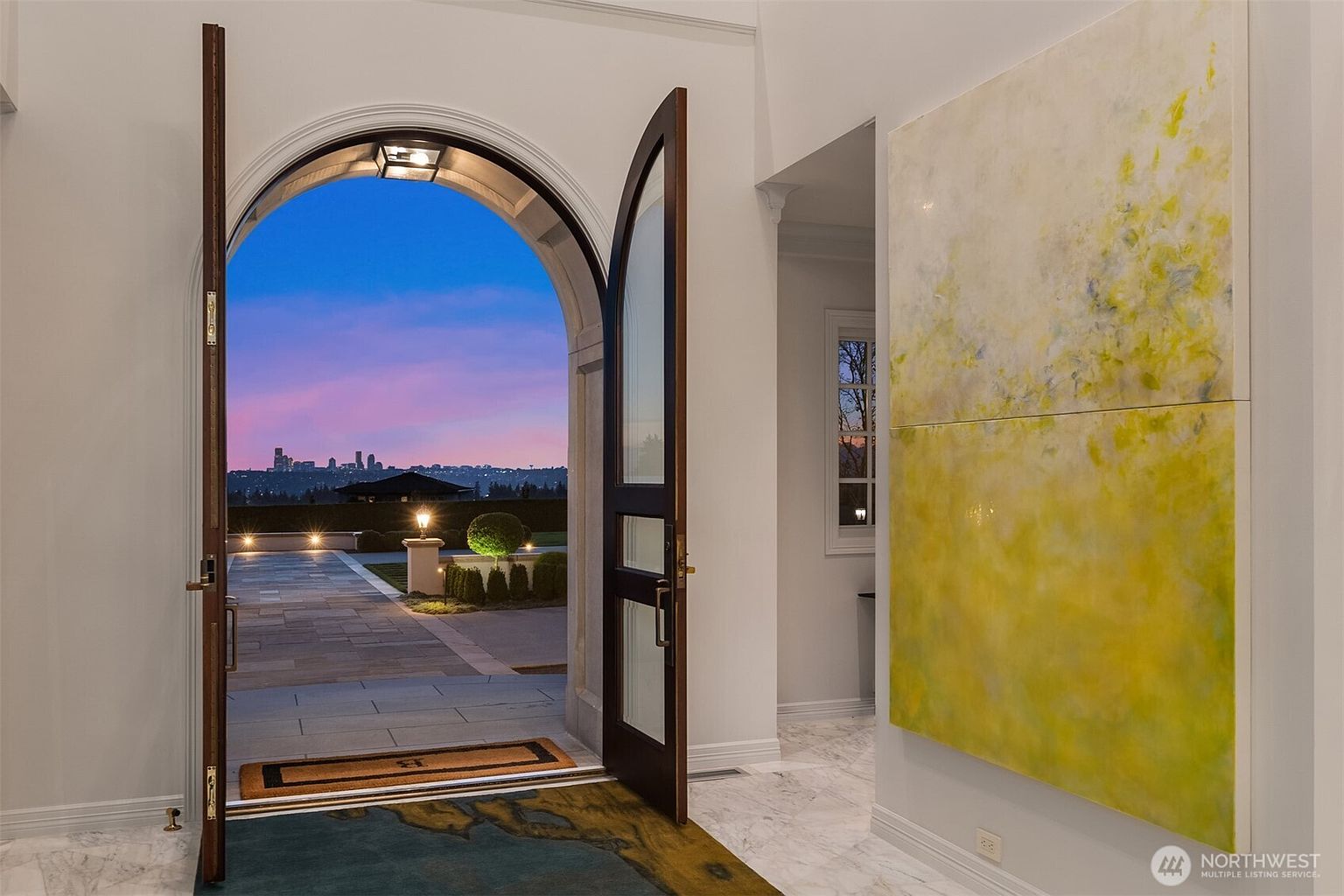 This elegant entryway features a grand arched doorway that opens to a stunning sunset view of a city skyline. The interior is characterized by polished marble flooring and a large, vibrant abstract painting on the wall, creating a sophisticated and welcoming atmosphere. The perspective captures the transition from the refined indoor space to the expansive outdoor patio, emphasizing the home's seamless connection to its scenic surroundings.