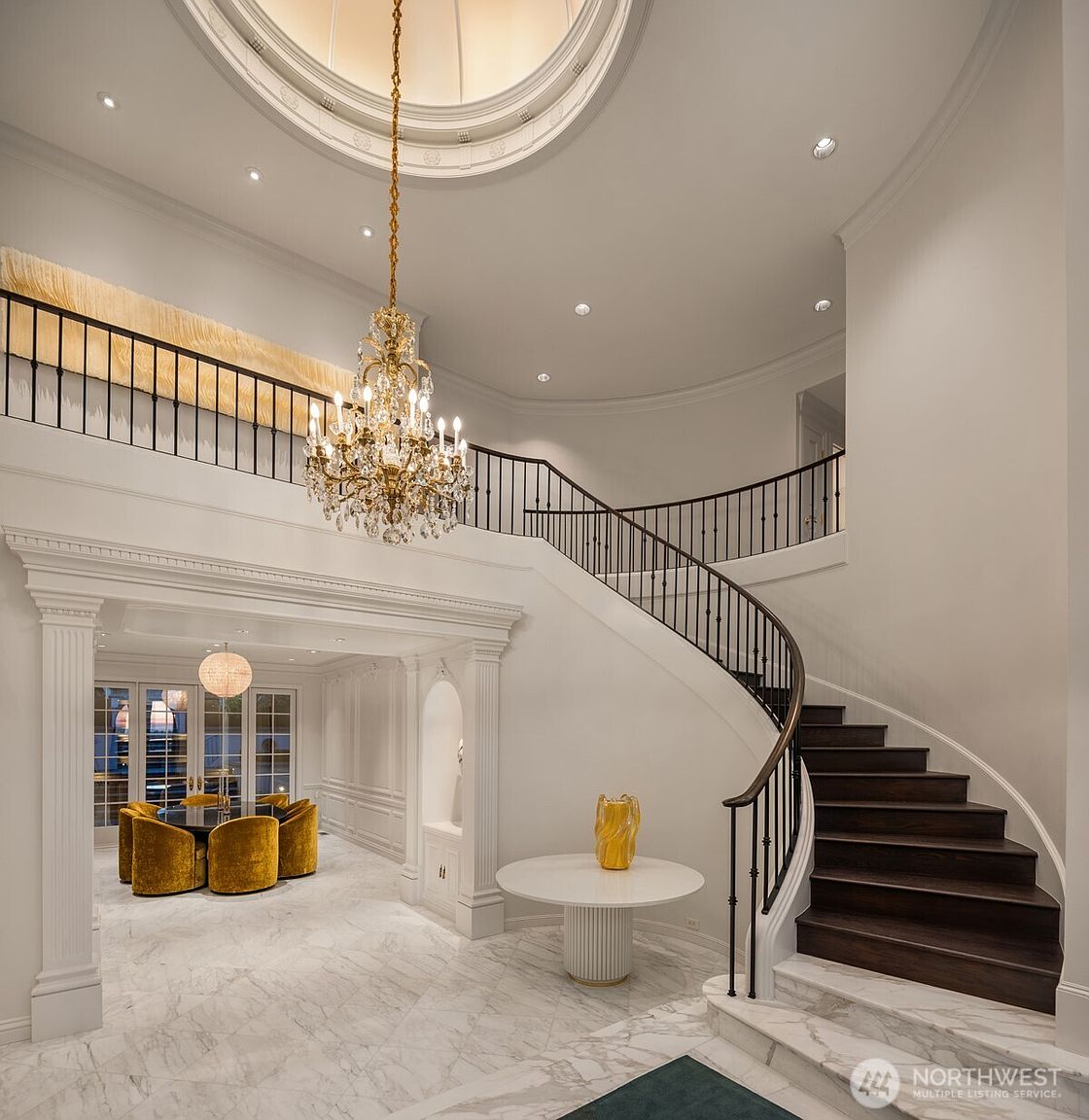 This grand foyer features a dramatic curved staircase with dark wood treads and a sleek black wrought-iron railing, set against a backdrop of pristine white walls and marble flooring. A magnificent crystal chandelier hangs from a recessed dome ceiling, casting a warm glow over the space, while a circular pedestal table with a gold vase serves as a sophisticated focal point. The open-concept design provides a clear view into an adjacent dining area, creating an atmosphere of luxury and architectural elegance.