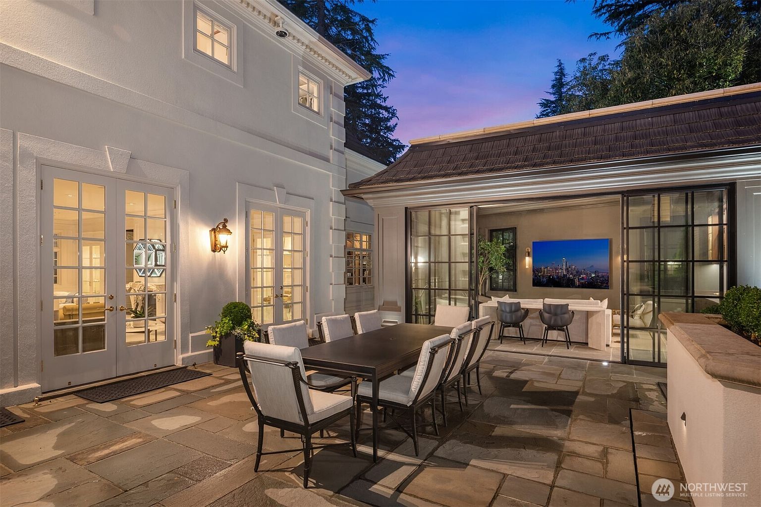 This elegant outdoor patio space features a stone-paved dining area with a large table and chairs, perfect for al fresco entertaining. The space seamlessly transitions into an indoor living area through expansive glass sliding doors, creating a sophisticated indoor-outdoor flow. The scene is set at dusk, with warm lighting illuminating the home's architectural details and the cozy interior lounge.