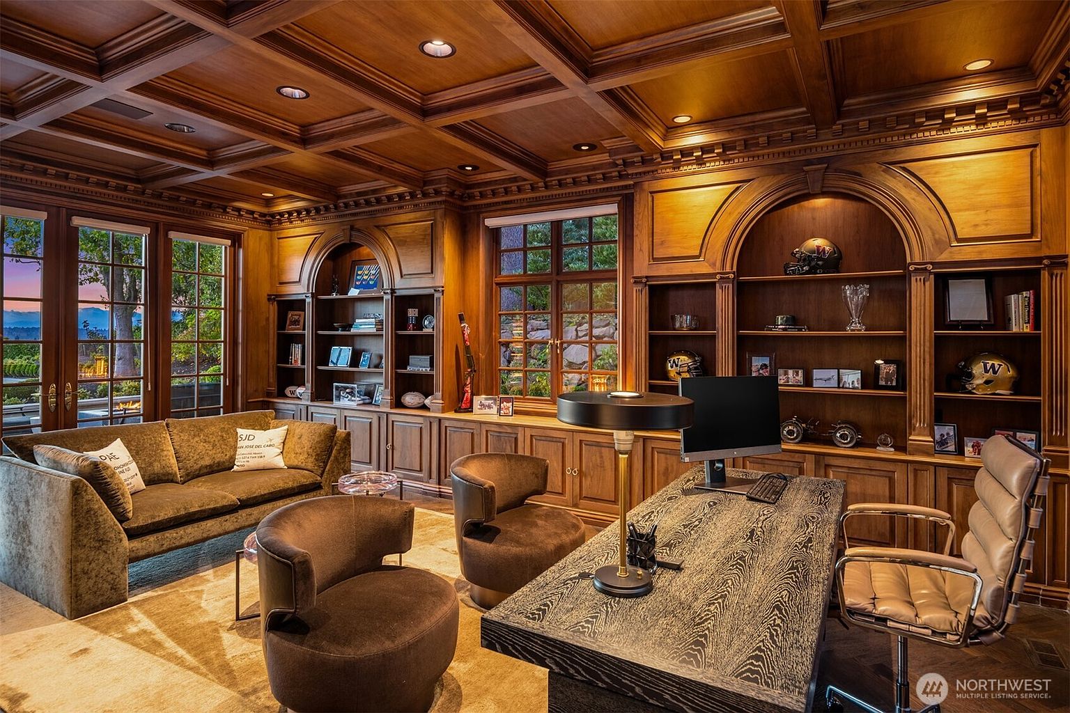 This sophisticated home office features rich, dark wood paneling, a coffered ceiling, and built-in shelving units that create a warm, scholarly atmosphere. The room is furnished with a textured wooden desk, a modern office chair, and a plush velvet sofa, all arranged to overlook a scenic outdoor view through large glass doors. The combination of classic architectural details and contemporary furniture makes this an ideal space for focused work or quiet reflection.