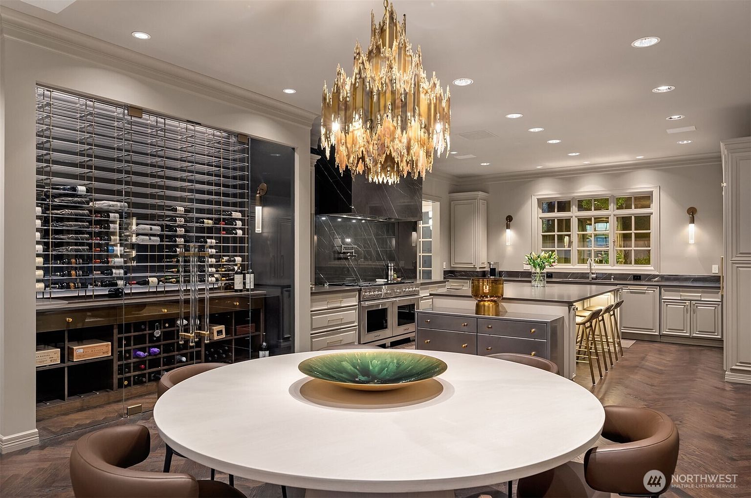 This luxurious, open-concept kitchen features a large central island with bar seating, a professional-grade range, and a sophisticated, climate-controlled glass wine cellar. A prominent, ornate gold chandelier hangs above a circular dining table in the foreground, creating a dramatic focal point. The space is defined by high-end finishes, dark cabinetry, and a seamless blend of modern elegance and functional design.