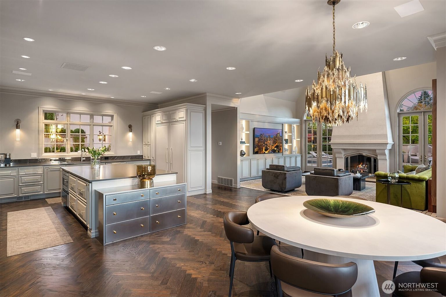 This expansive, open-concept kitchen and living area features a sophisticated blend of traditional and modern design, highlighted by dark herringbone hardwood floors and a striking metallic-faced kitchen island. A grand, ornate chandelier hangs above a circular dining table in the foreground, while the background showcases a cozy living space with a fireplace and built-in media cabinetry. The room is bathed in natural light from large windows and glass doors, creating an elegant and inviting atmosphere perfect for entertaining.
