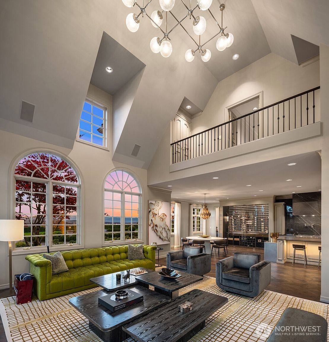 This expansive, high-ceilinged living room features a striking lime green tufted sofa, dark wood coffee tables, and plush grey armchairs, creating a sophisticated yet vibrant atmosphere. The space is anchored by a grand chandelier and large arched windows that frame a scenic sunset view, while an open-concept layout flows seamlessly into a dining area and a modern kitchen with a wine cellar. The architectural design includes a mezzanine balcony with a black iron railing, emphasizing the home's airy, luxurious, and open-plan aesthetic.