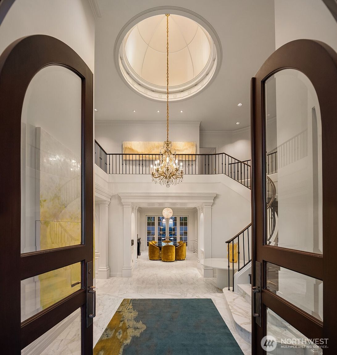 This grand entryway features a dramatic double-height foyer with a sweeping marble staircase and a stunning crystal chandelier suspended from a circular domed ceiling. The space is finished with polished marble flooring, a vibrant area rug, and elegant architectural molding, creating a sophisticated and luxurious first impression. The perspective looks through arched glass-paneled doors into a bright, open-concept home with a glimpse of a formal dining area in the background.