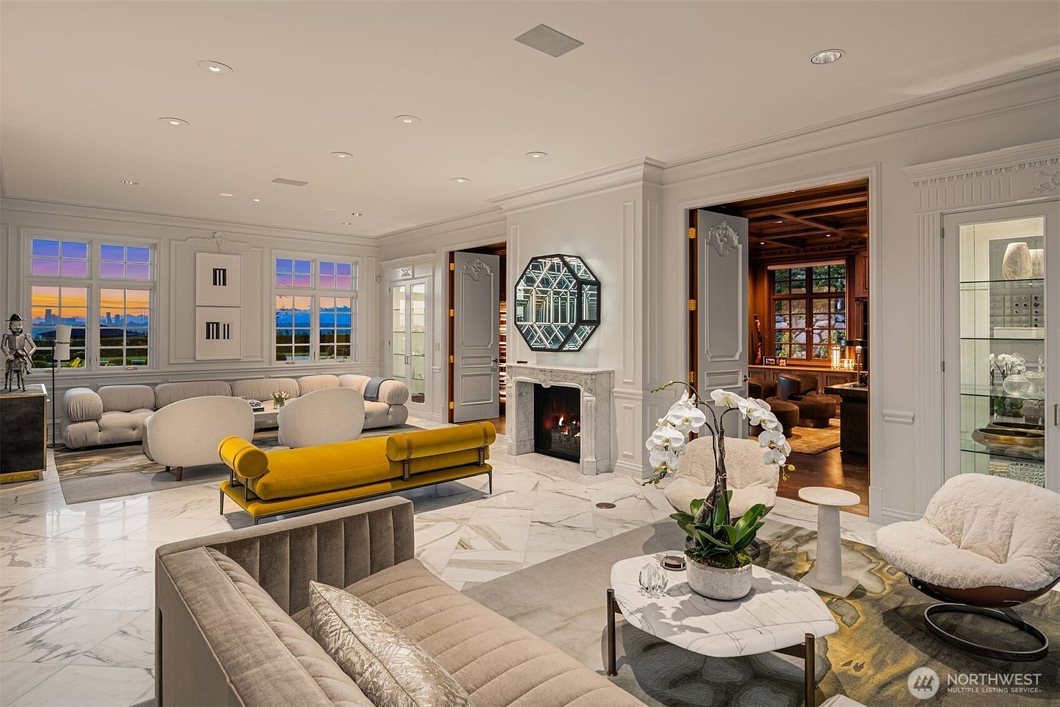 This expansive, luxurious living room features a sophisticated blend of modern and classic design elements, including polished marble flooring and intricate wall molding. The space is anchored by a central fireplace with a striking geometric mirror above, flanked by a vibrant yellow velvet daybed and plush, neutral-toned seating arrangements. Large windows offer a picturesque sunset view, while an adjacent wood-paneled study adds a warm, rich contrast to the bright, airy main living area.