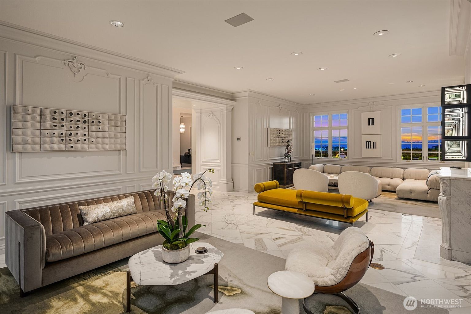 This expansive, luxurious living room features a sophisticated blend of classic architectural molding and contemporary furnishings, set against a backdrop of polished marble flooring. The space is anchored by a plush velvet sofa and a vibrant mustard-yellow daybed, while large windows offer a stunning sunset view of the ocean. The room exudes an air of refined elegance, enhanced by curated art pieces and a neutral color palette that highlights the high-end finishes.