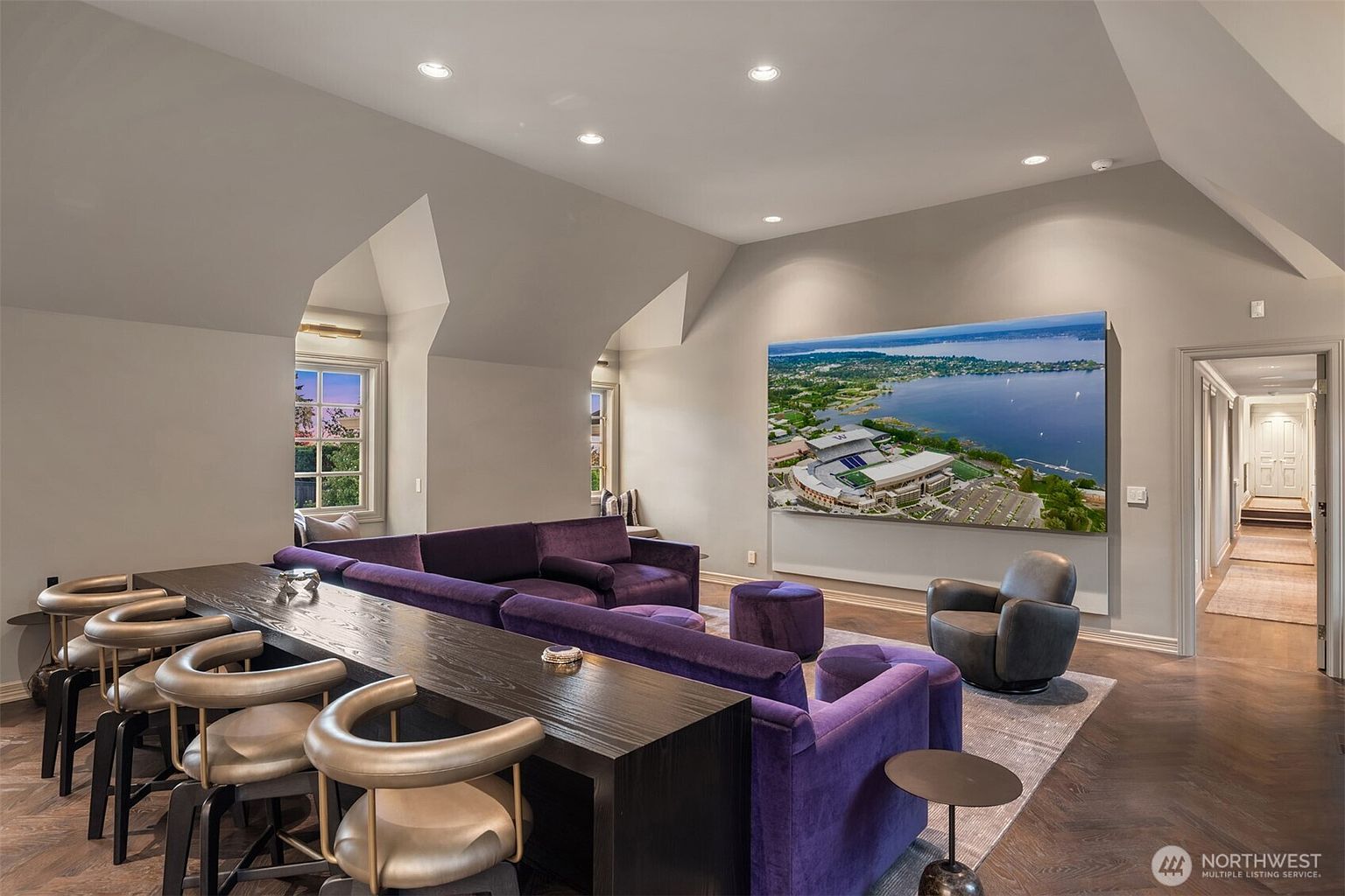 This spacious and sophisticated media room features a large, plush purple sectional sofa and a sleek, dark wood bar table with matching metallic stools. A massive wall-mounted screen displays an aerial view of a stadium, serving as the room's focal point, while recessed lighting and vaulted ceilings enhance the airy, modern atmosphere. The room is designed for comfort and entertainment, seamlessly blending luxury seating with functional social spaces.