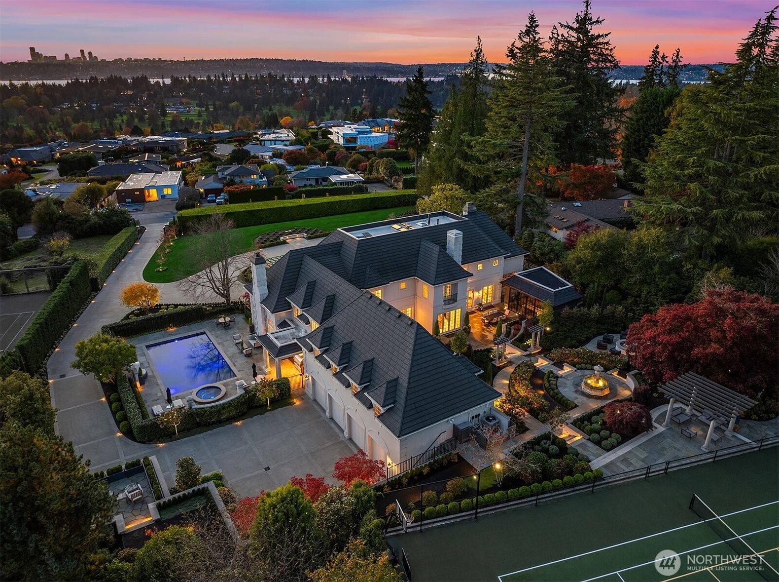 This stunning aerial view captures a luxurious estate at dusk, showcasing a sprawling residence with a dark-tiled roof, a pristine swimming pool with an integrated spa, and a private tennis court. The meticulously landscaped grounds feature manicured gardens, stone pathways, and a decorative fountain, all set against a breathtaking sunset backdrop with a distant city skyline. The perspective highlights the property's expansive layout and high-end outdoor amenities, emphasizing its exclusivity and sophisticated design.