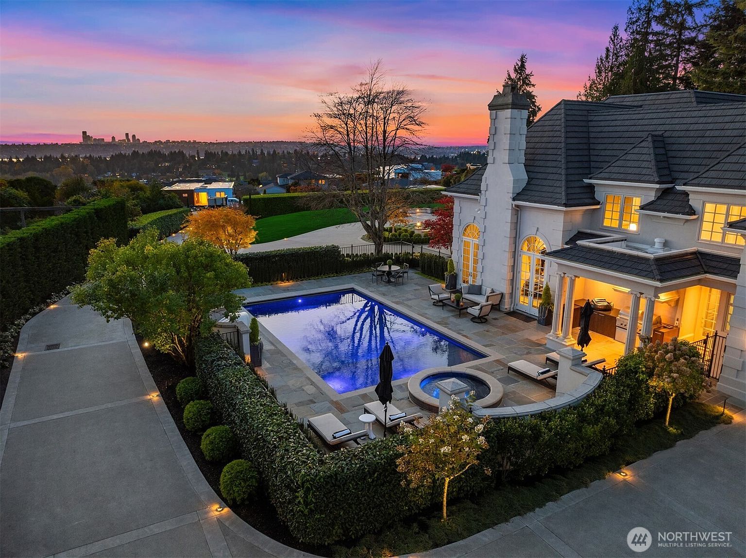 This stunning aerial view captures a luxurious estate at sunset, featuring a pristine rectangular swimming pool and integrated spa set within a beautifully landscaped stone patio. The elegant home showcases classic architectural details, while the background offers a breathtaking panoramic view of a distant city skyline under a vibrant, colorful sky. The scene exudes an atmosphere of high-end sophistication and tranquil outdoor living.