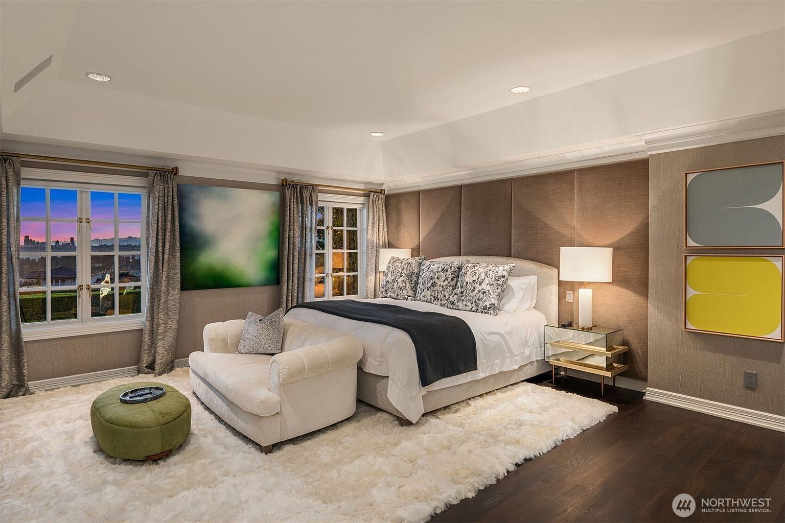 This luxurious primary bedroom features a sophisticated design with a large, upholstered bed as the focal point, complemented by a plush white area rug and a cozy cream-colored chaise lounge. The room is accented by a textured, paneled headboard wall, modern art pieces, and elegant mirrored nightstands, all set against dark hardwood floors. Large windows with soft drapery allow for a beautiful sunset view, creating a serene and high-end atmosphere.