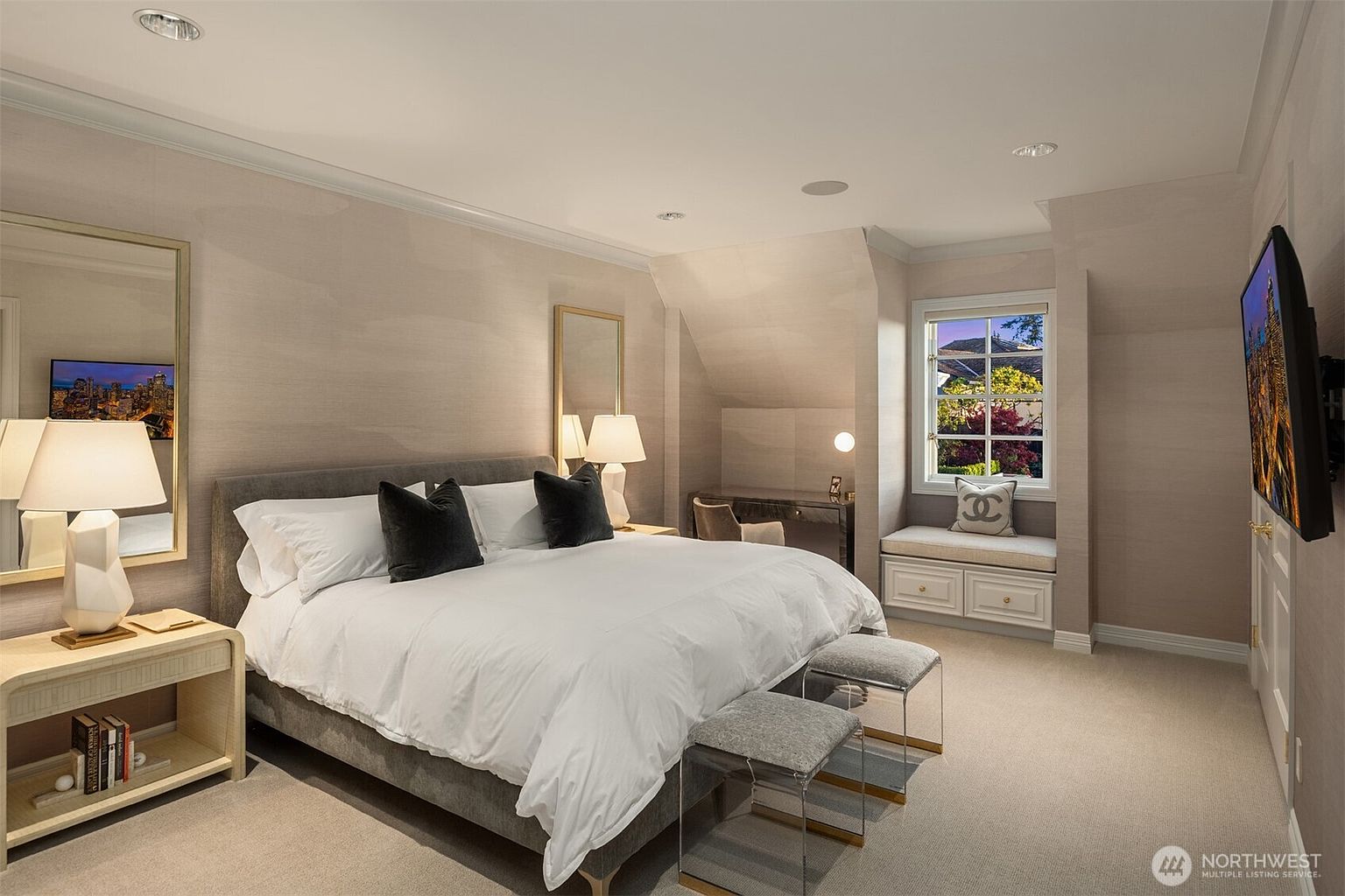 This elegant primary bedroom features a plush king-sized bed with a grey upholstered headboard, accented by dark decorative pillows and crisp white linens. The room is bathed in soft, neutral tones, complemented by a built-in window seat, a sleek desk area, and two modern acrylic-based benches at the foot of the bed. The space exudes a sophisticated, tranquil atmosphere, enhanced by ambient lighting and a large wall-mounted television.