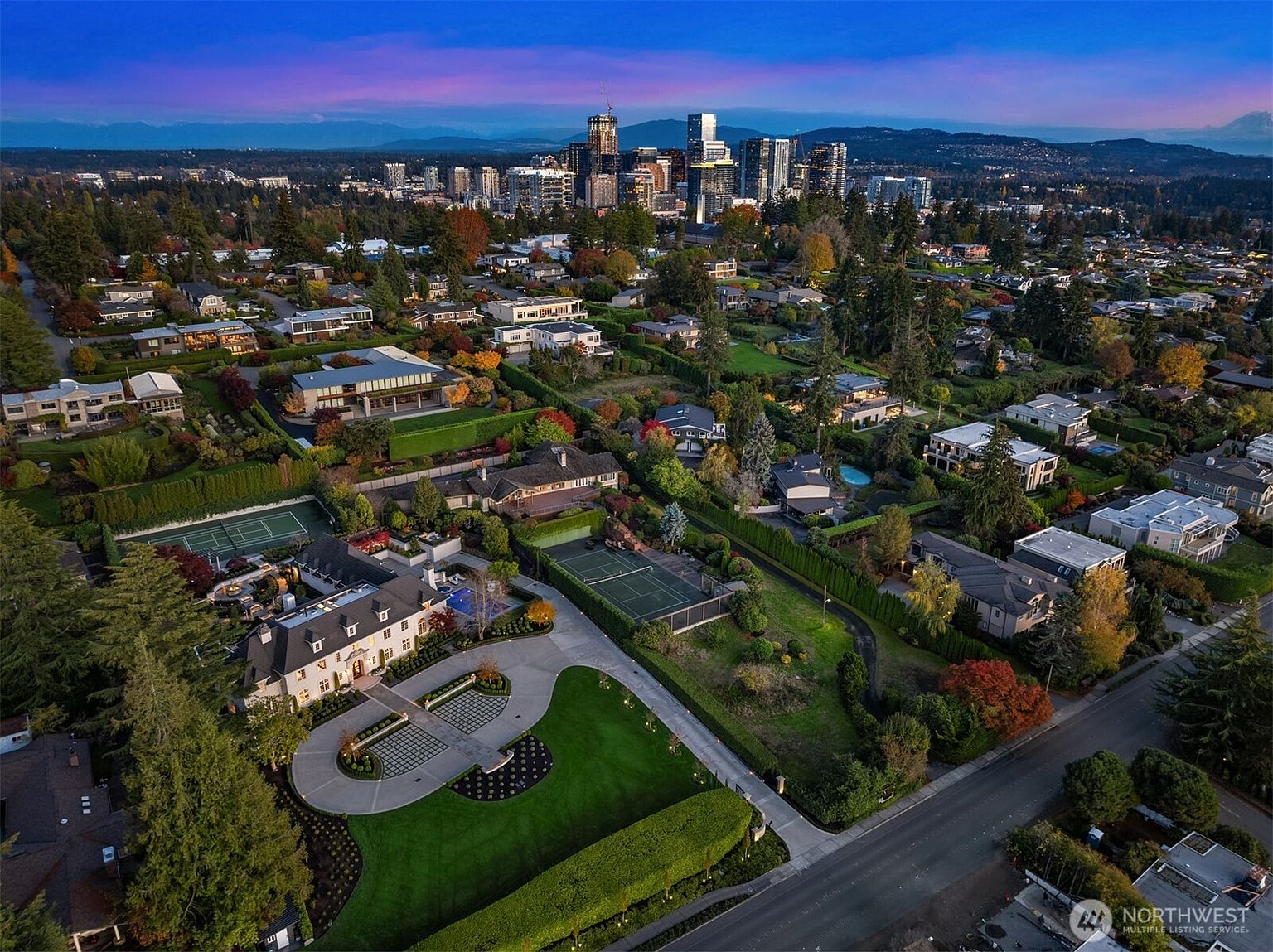 This stunning aerial view captures a sprawling luxury estate in a lush, tree-lined neighborhood, featuring a grand mansion with a private tennis court and manicured grounds. The perspective showcases the property's expansive layout in relation to the vibrant city skyline and distant mountain range during a picturesque twilight. The overall impression is one of exclusivity, prestige, and high-end suburban living.