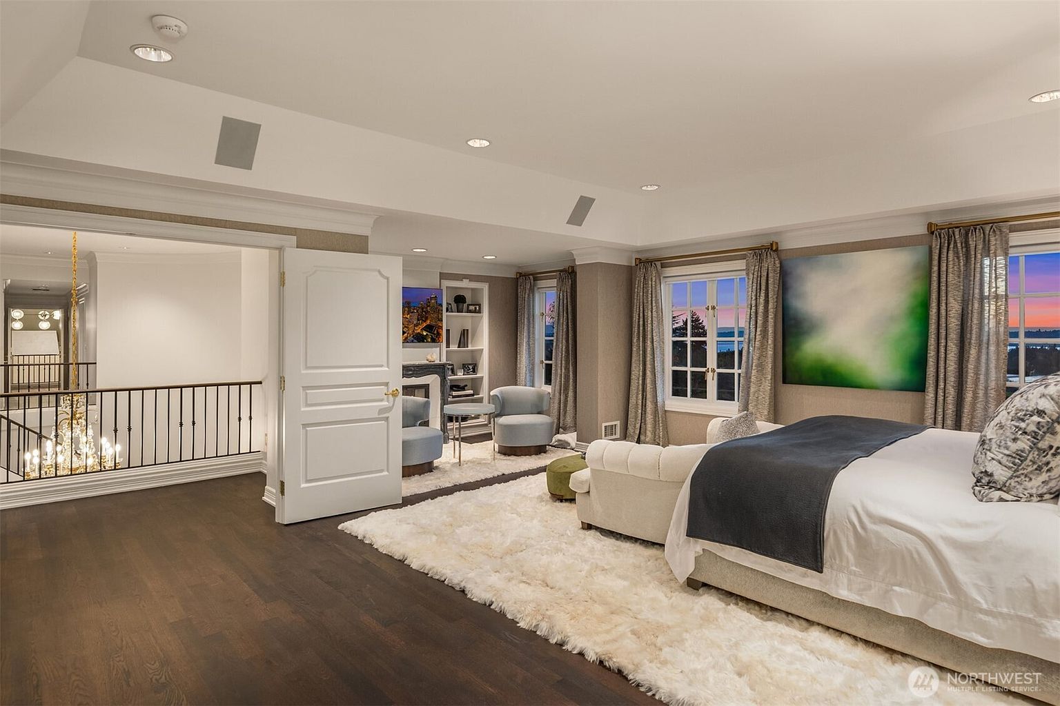 This luxurious primary bedroom features a plush, cream-colored tufted bed as the centerpiece, set against a backdrop of soft neutral walls and a large, vibrant abstract painting. The room includes a cozy seating area near a fireplace and built-in shelving, with expansive windows that offer a scenic view of the water at dusk. A dark hardwood floor transitions into a thick, textured area rug, creating a sophisticated and tranquil atmosphere.