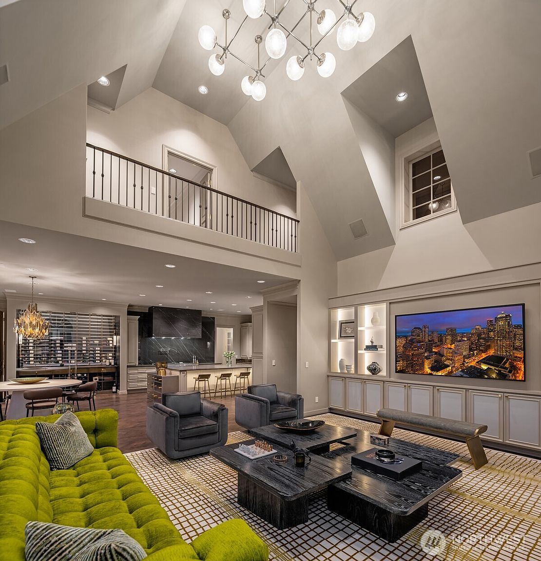 This expansive, high-ceilinged living room features a striking lime green tufted sofa, dark charcoal armchairs, and a multi-tiered modern chandelier that serves as a focal point. The open-concept layout seamlessly transitions into a sophisticated kitchen area, while a built-in media wall with integrated shelving and a large television anchors the space. The room exudes a luxurious, contemporary aesthetic with a dramatic sense of scale and refined architectural details.