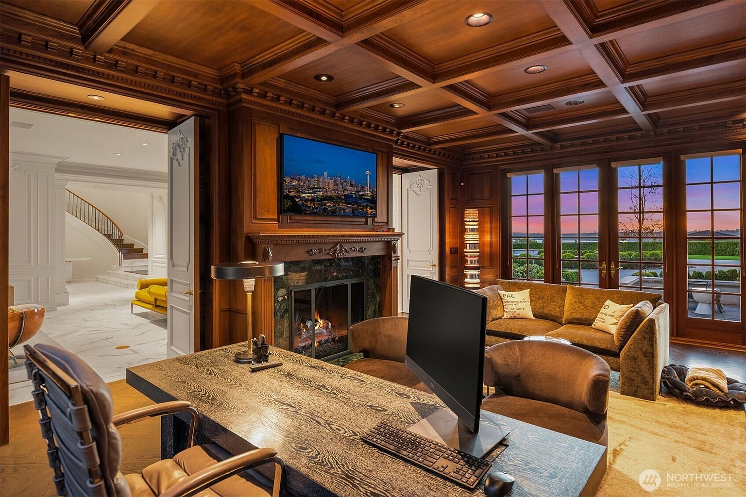 This sophisticated home office exudes luxury with its rich, dark wood-paneled walls and an intricate coffered ceiling. The room features a large, textured executive desk, a cozy seating area with a plush sofa, and a fireplace, all framed by expansive windows that offer a stunning sunset view. The perspective captures the professional yet inviting atmosphere, seamlessly connecting to a bright, marble-floored hallway with a grand staircase in the background.